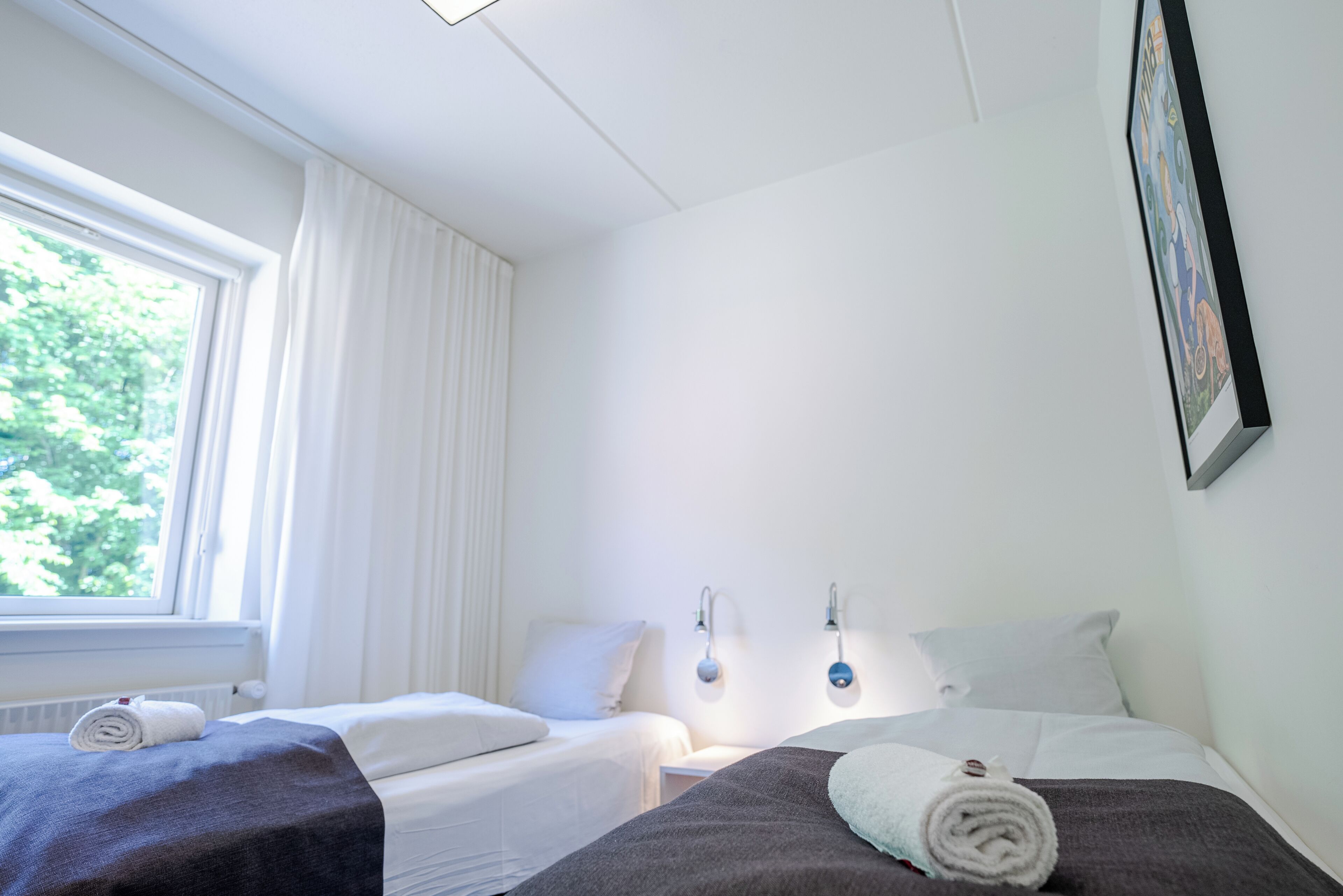 Habitación de hotel con dos camas, una ventana con cortinas, un cuadro colgado en la pared y una toalla sobre una de las camas.