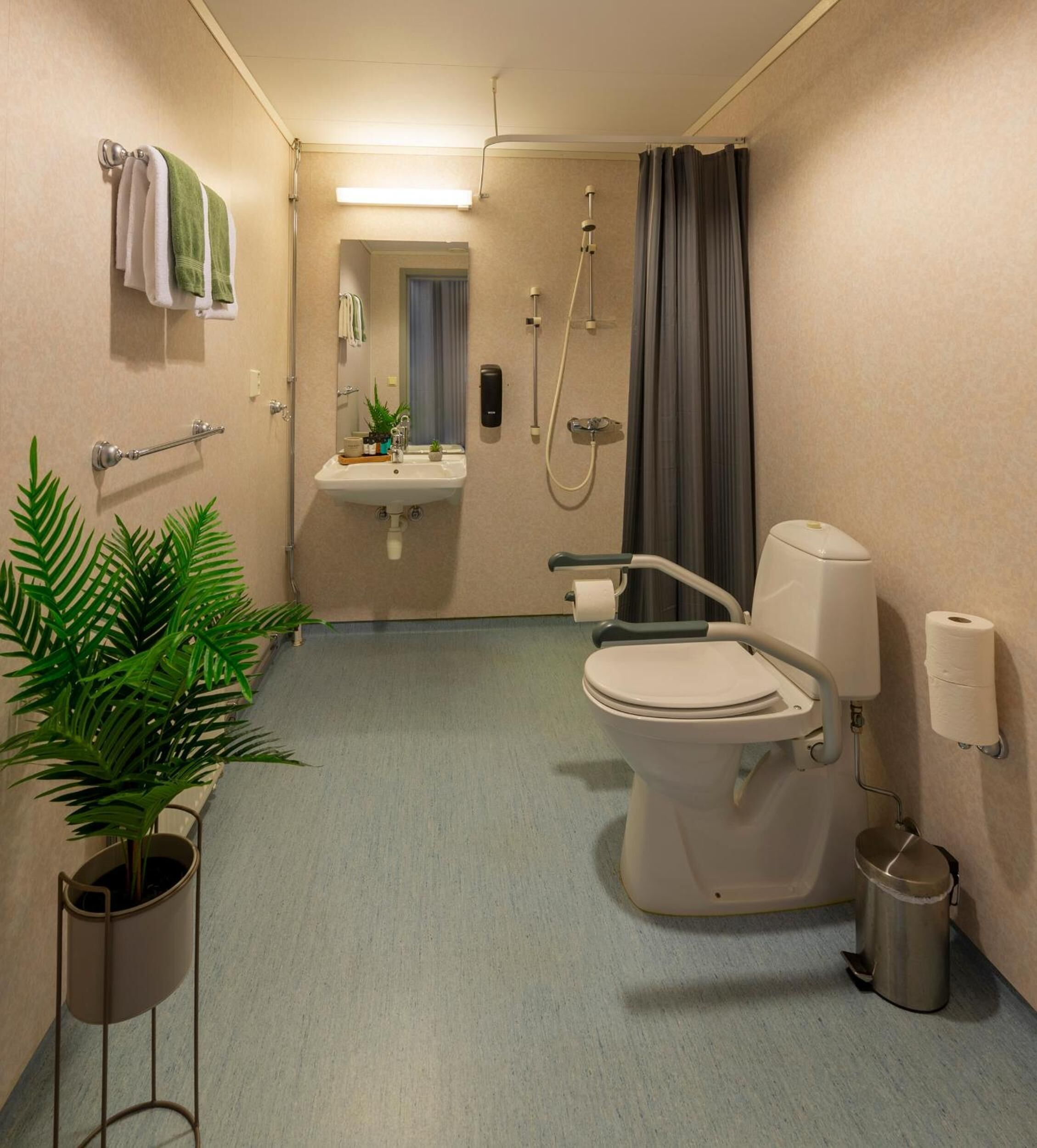 Une petite salle de bain comprenant des toilettes, un lavabo et une douche. On y trouve des serviettes et une plante accrochée au mur, ainsi qu'une poubelle.