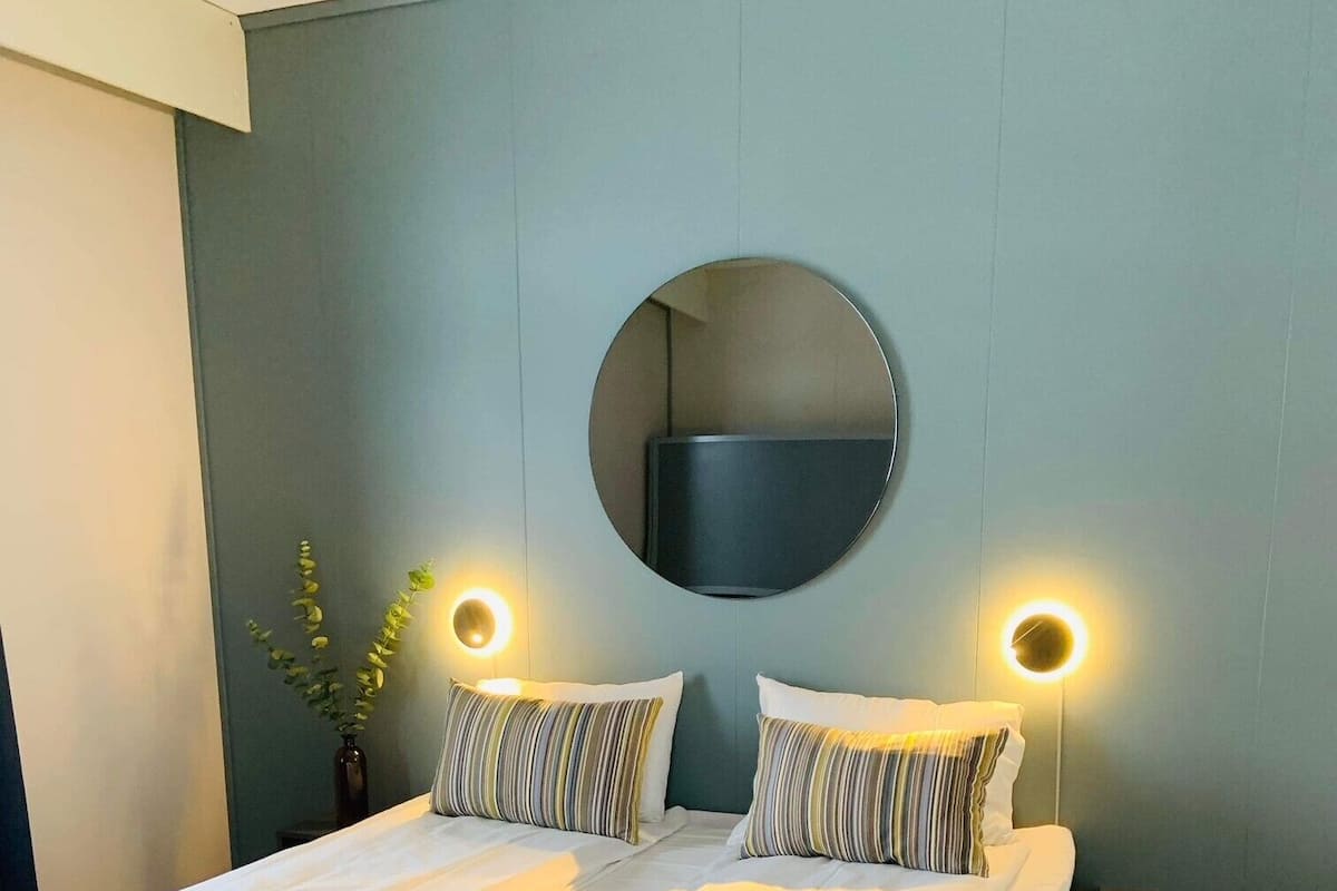 A modern bedroom with a large bed, two bedside tables, a round mirror, and wall-mounted lights.