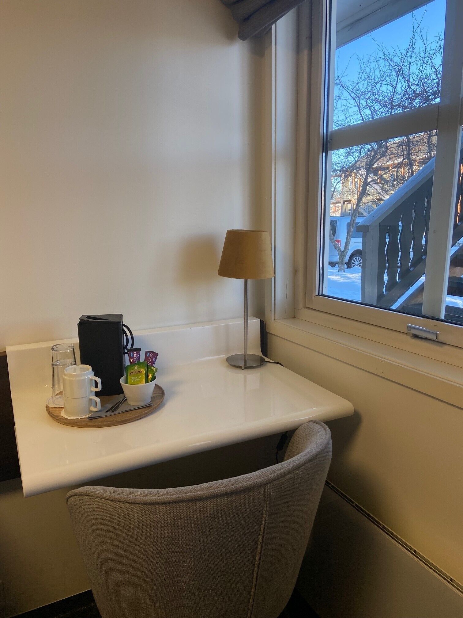 A corner of a room with a white table, a lamp, a chair, and a window with a view of a building and trees.