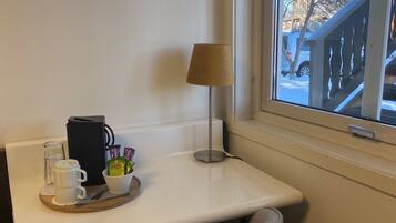 A corner of a room with a white table, a lamp, a chair, and a window with a view of a building and trees.