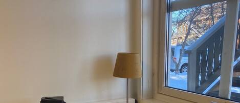 Un rincón de una habitación con una mesa blanca, una lámpara, una silla y una ventana que da a un edificio y árboles.