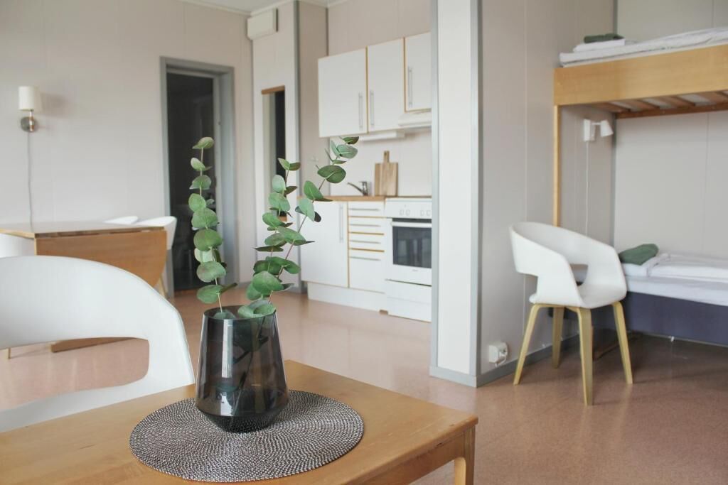 Ein modernes Zimmer mit einem Holztisch, einem weißen Stuhl, einer kleinen Topfpflanze, einem Etagenbett und einer Küchenzeile im Hintergrund.