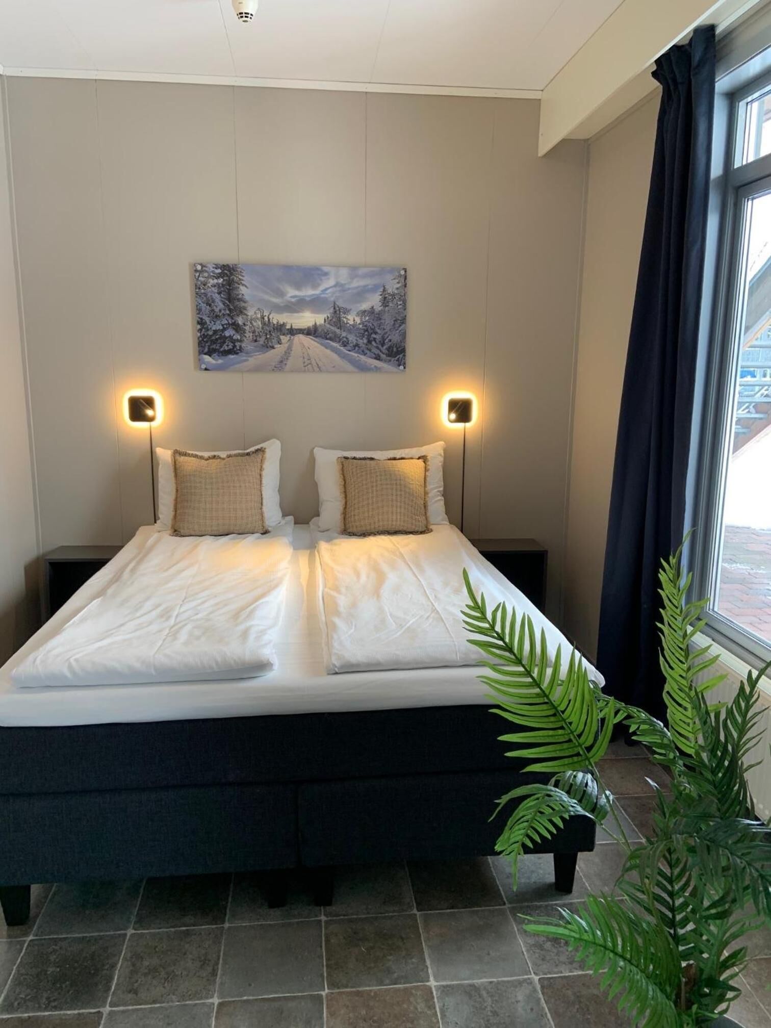 A double bed with white linens, two bedside lamps, a painting of a snowy landscape, and a potted plant.
