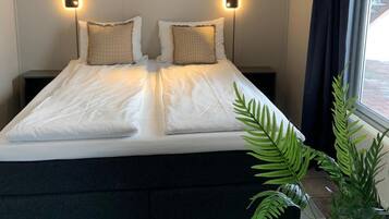 A double bed with white linens, two bedside lamps, a painting of a snowy landscape, and a potted plant.