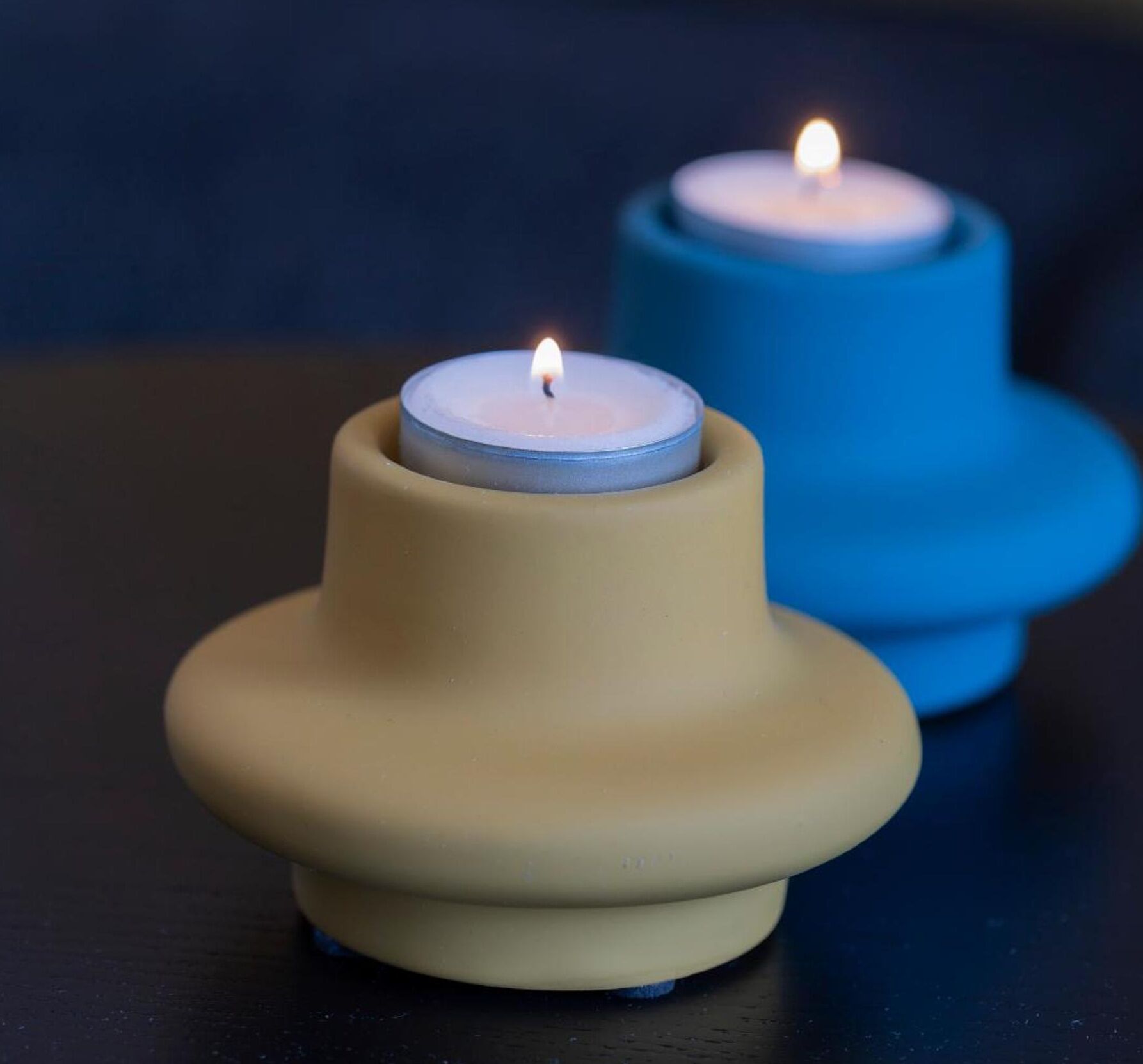 Duas velas em castiçais, uma bege e outra azul, sobre uma superfície escura.