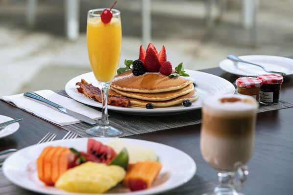 Desayuno con tortitas, fruta y bebidas sobre una mesa.