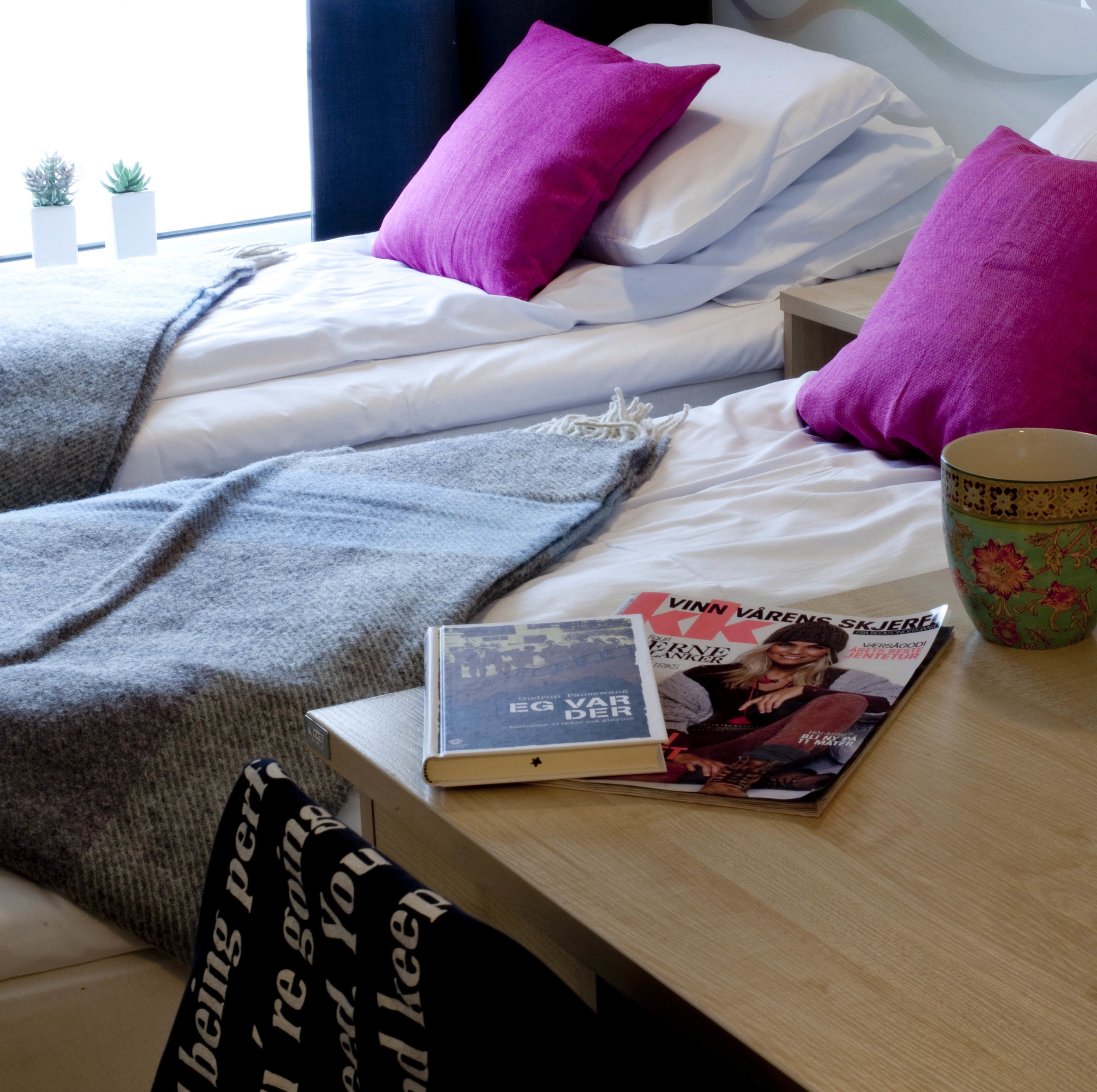 A neatly made bed with white and purple pillows, a grey blanket, and a magazine on a wooden bedside table.