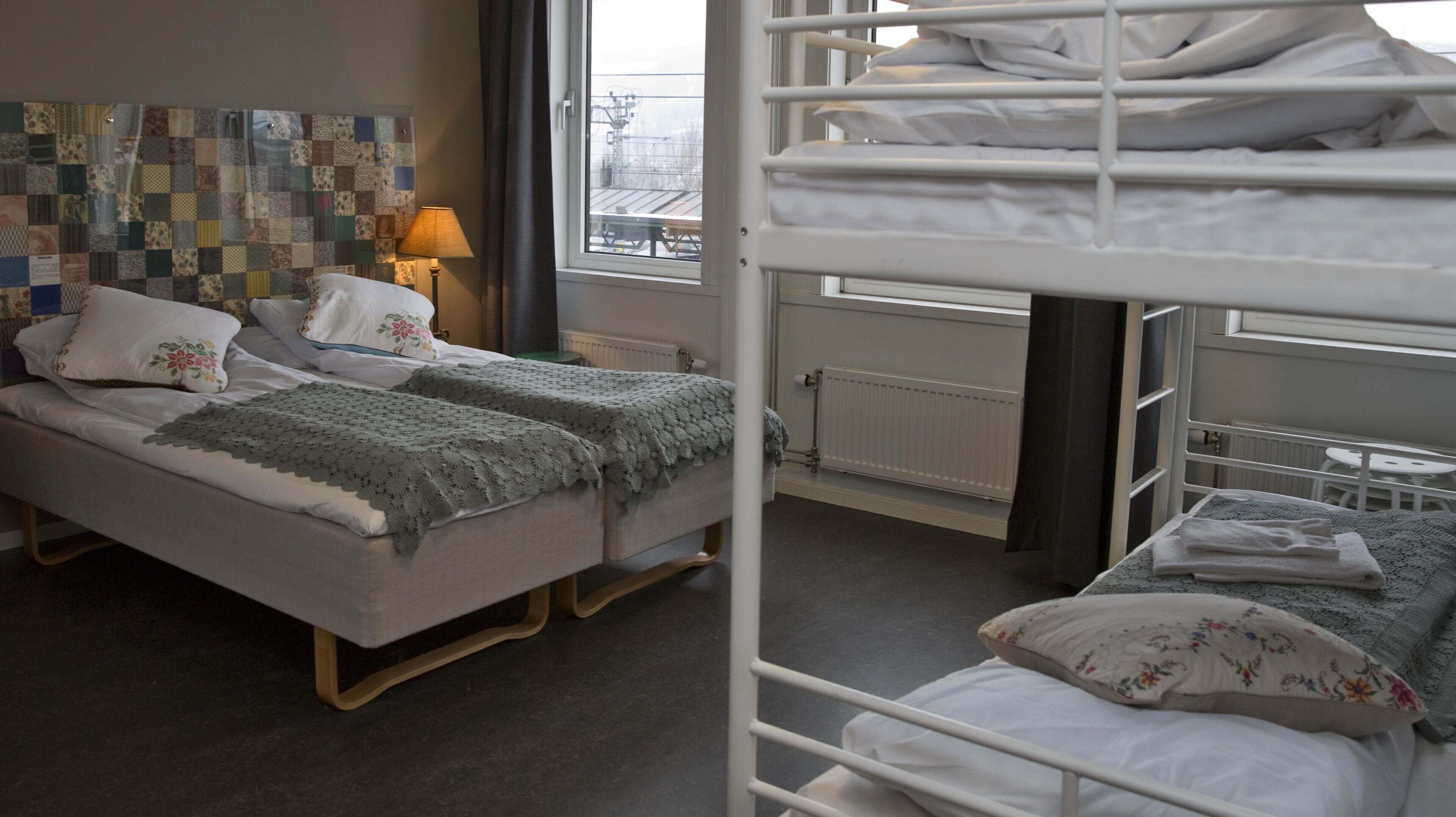 Habitación de hotel con una cama individual, una cama adicional en literas, un cabecero acolchado, una lámpara y una ventana con vistas al exterior.