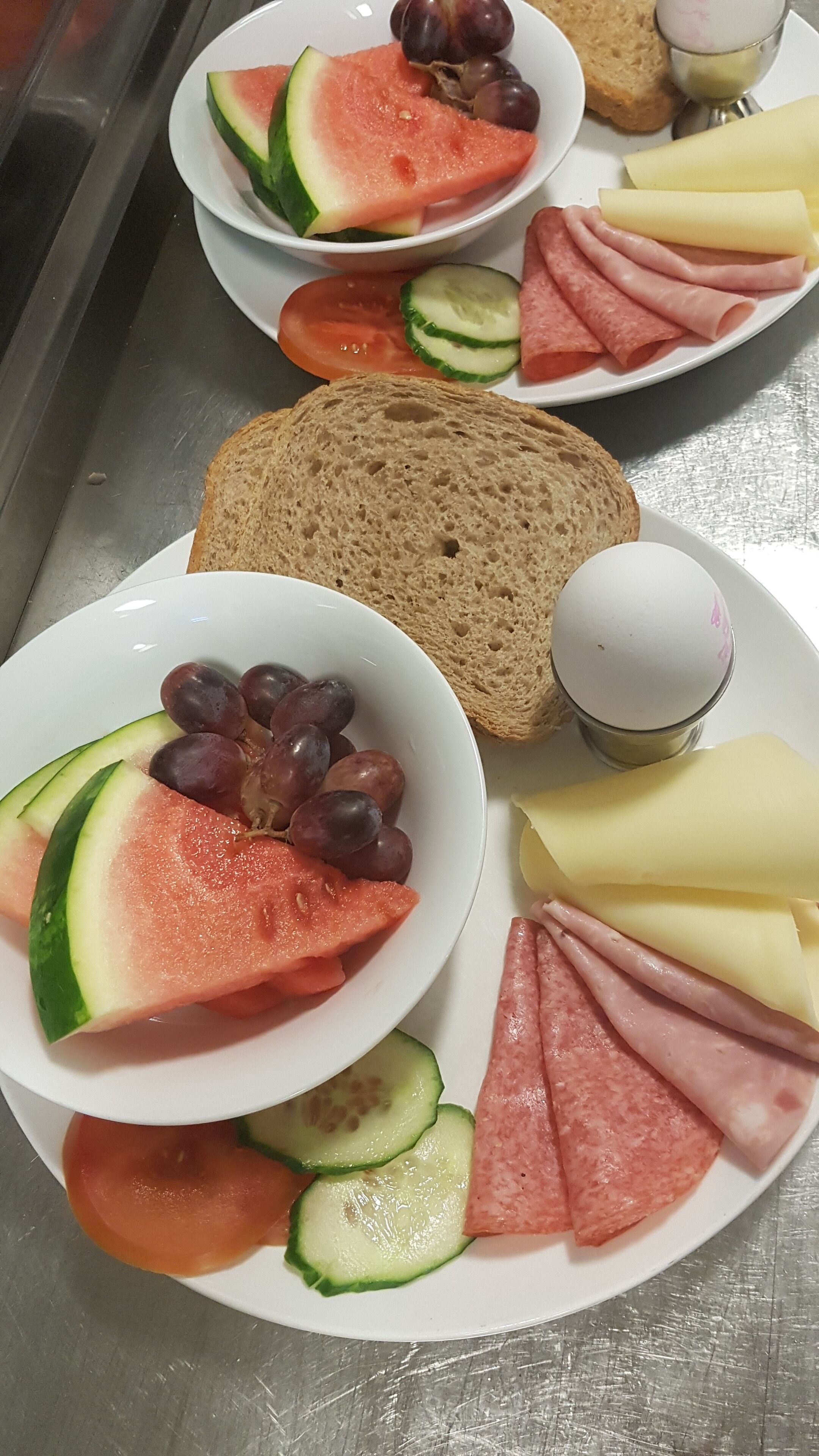 A meal with watermelon, grapes, cucumber slices, ham, cheese, and bread on a stainless steel table.