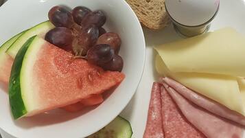 A meal with watermelon, grapes, cucumber slices, ham, cheese, and bread on a stainless steel table.