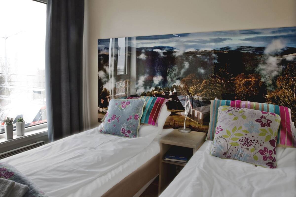 Quarto de hotel com duas camas, uma janela grande, vista para uma paisagem e uma mesa de cabeceira com abajur.