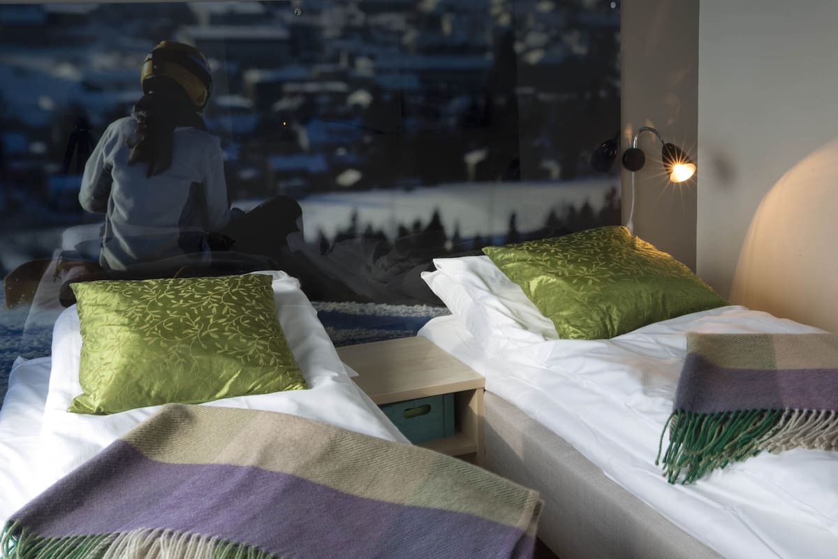 Quarto de hotel com duas camas, cada uma com um travesseiro e um cobertor verdes. Há uma mesa de cabeceira com uma luminária entre as camas.