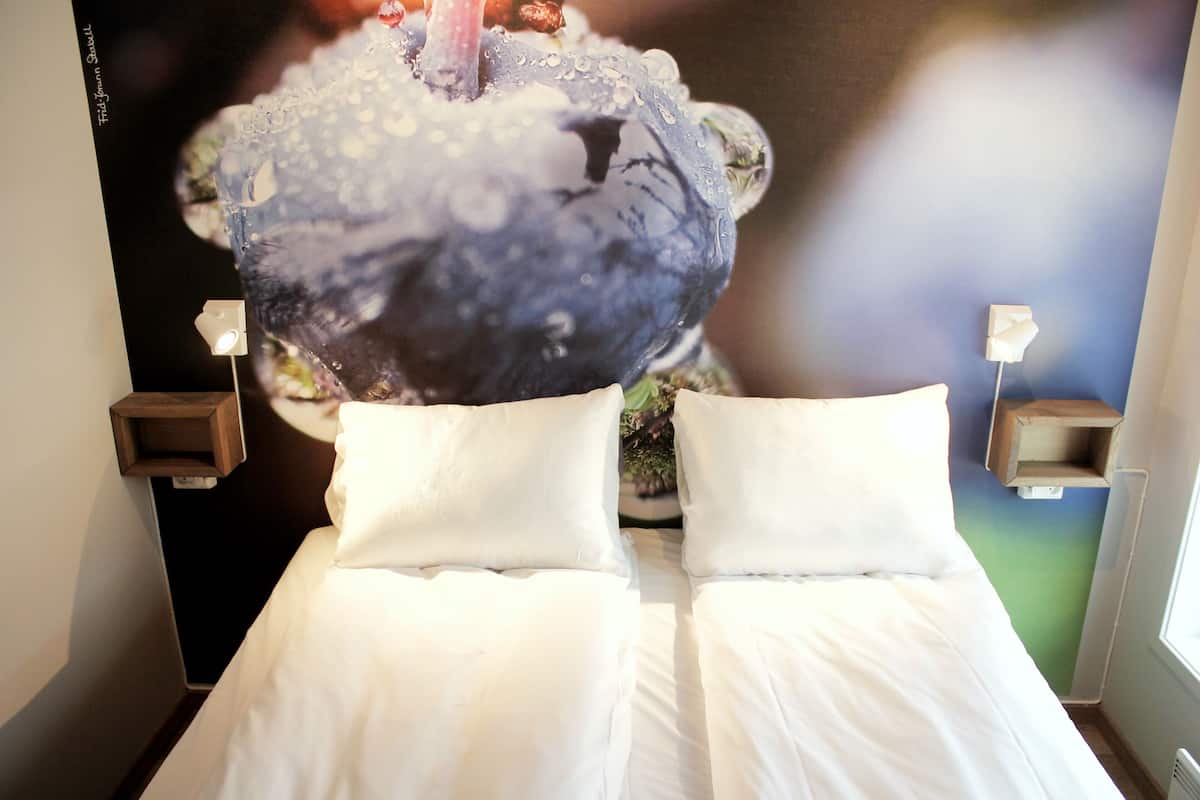 Cama com lençóis brancos e duas almofadas, mesa de cabeceira fixada na parede com abajur, e pôster de um objeto azul com gotas de água.