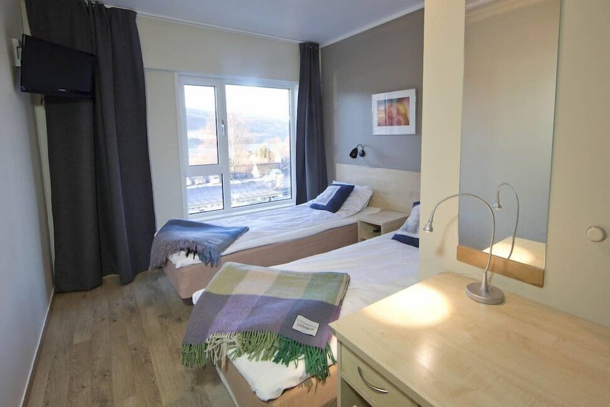 Quarto de hotel com duas camas, uma janela grande, televisão, mesa de cabeceira com abajur e espelho.