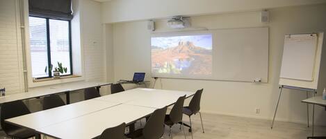 Un salón de conferencias con una larga mesa, sillas, una pantalla de proyector, una pizarra y una ventana con persianas.