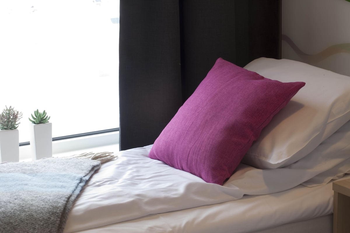 Cama bem arrumada com lençóis brancos e um único travesseiro roxo, uma plantinha na mesinha de cabeceira e uma janela com cortinas.