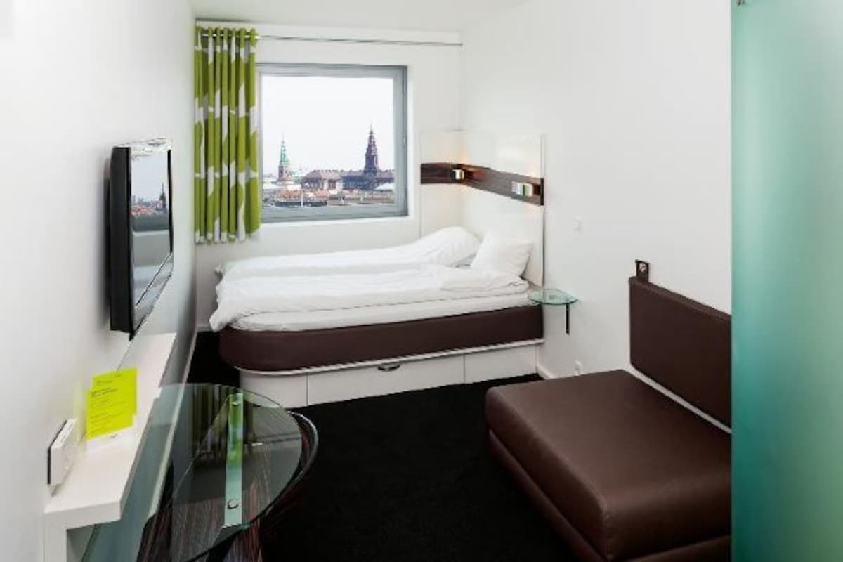 A hotel room with a bed, a desk, a TV, and a window with a view of a building.