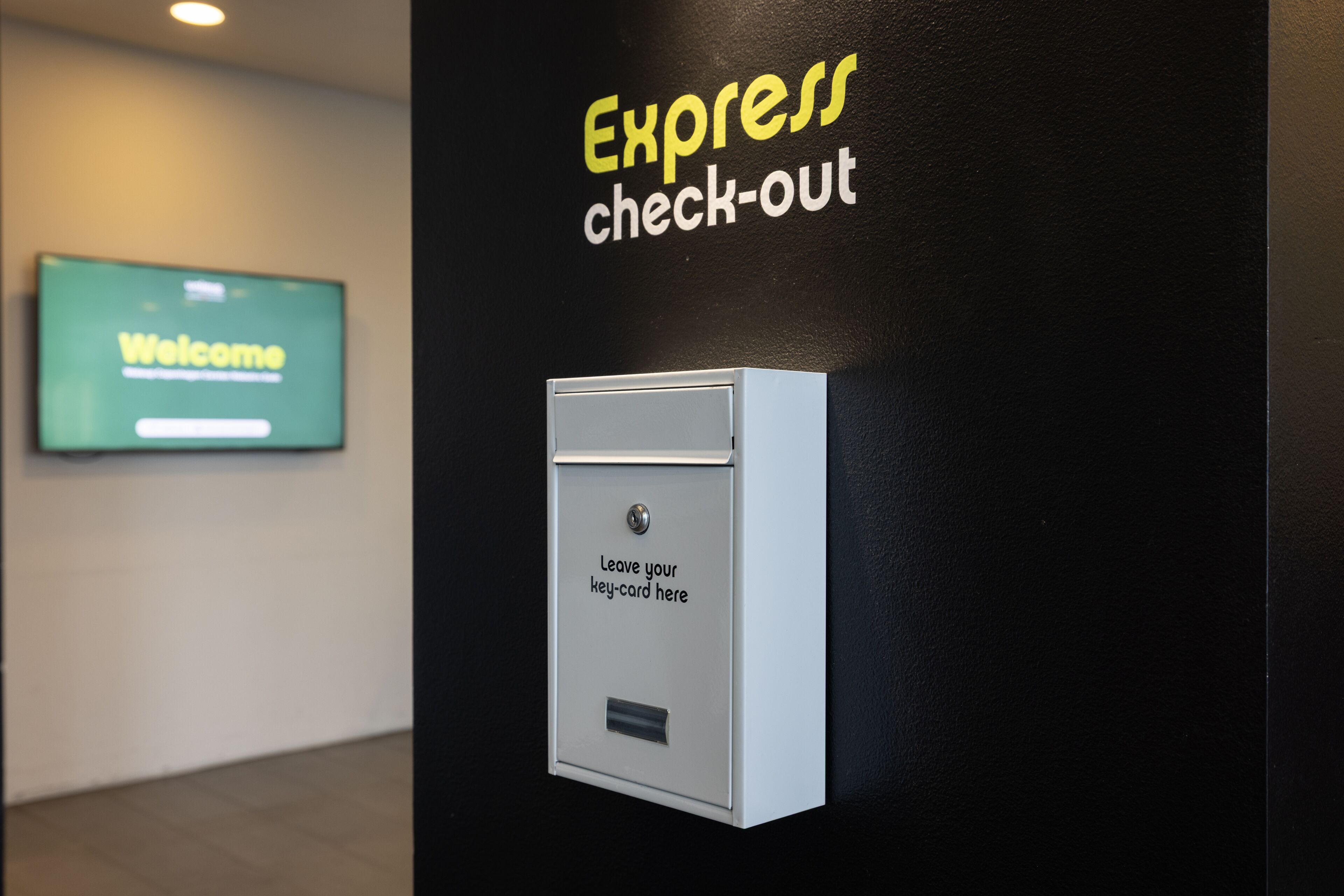 A wall-mounted electronic keypad with a sign above it reading "Express check-out" and a television screen in the background displaying "Welcome".