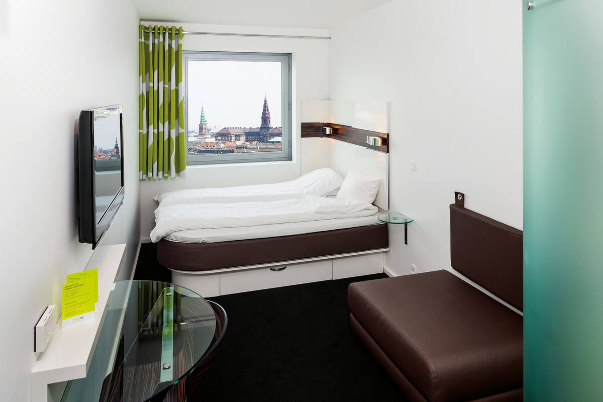 A compact hotel room with a bed, a small desk with a mirror, a glass table, a brown sofa, and a window offering a city view.