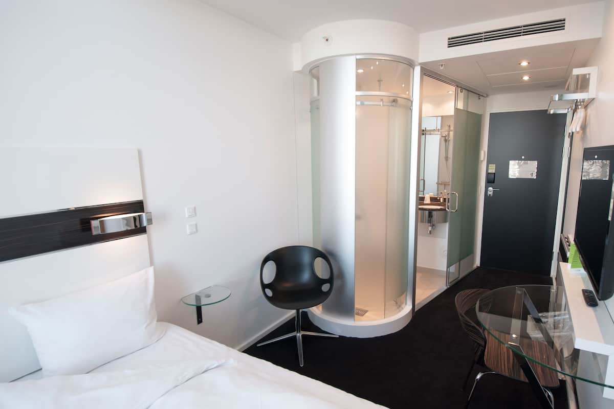 A modern hotel room with a bed, a glass-enclosed shower, a small table, and a chair.