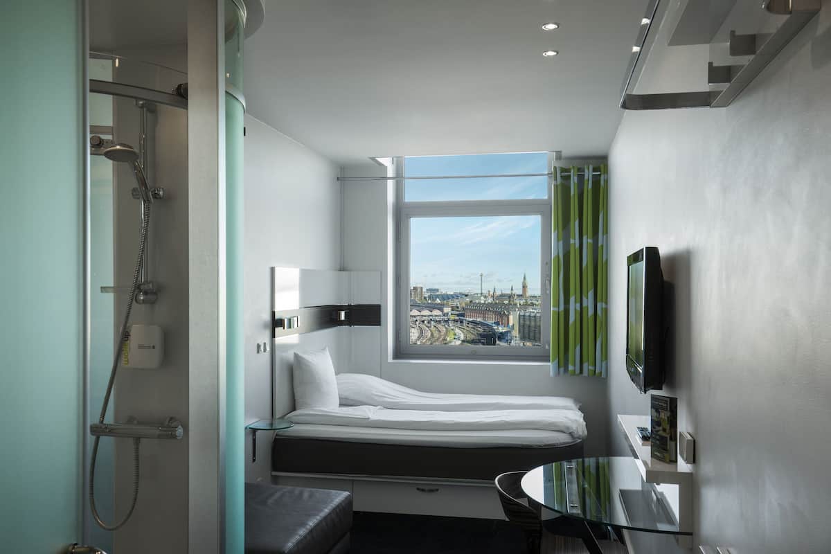 A modern hotel room with a bed, a glass table, a TV, and a shower.