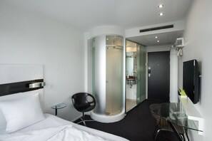 A modern hotel room with a bed, a glass shower enclosure, a glass table, a black chair, and a TV mounted on the wall. - Wakeup Copenhagen Carsten Niebuhrs Gade (Copenhagen)
