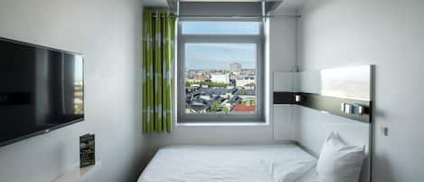 Habitación de hotel con cama, televisor, ventana con cortinas y vista a la ciudad.