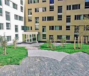 A courtyard with a paved walkway, a small garden area with young trees, and a multi-story building with a beige facade and numerous windows.