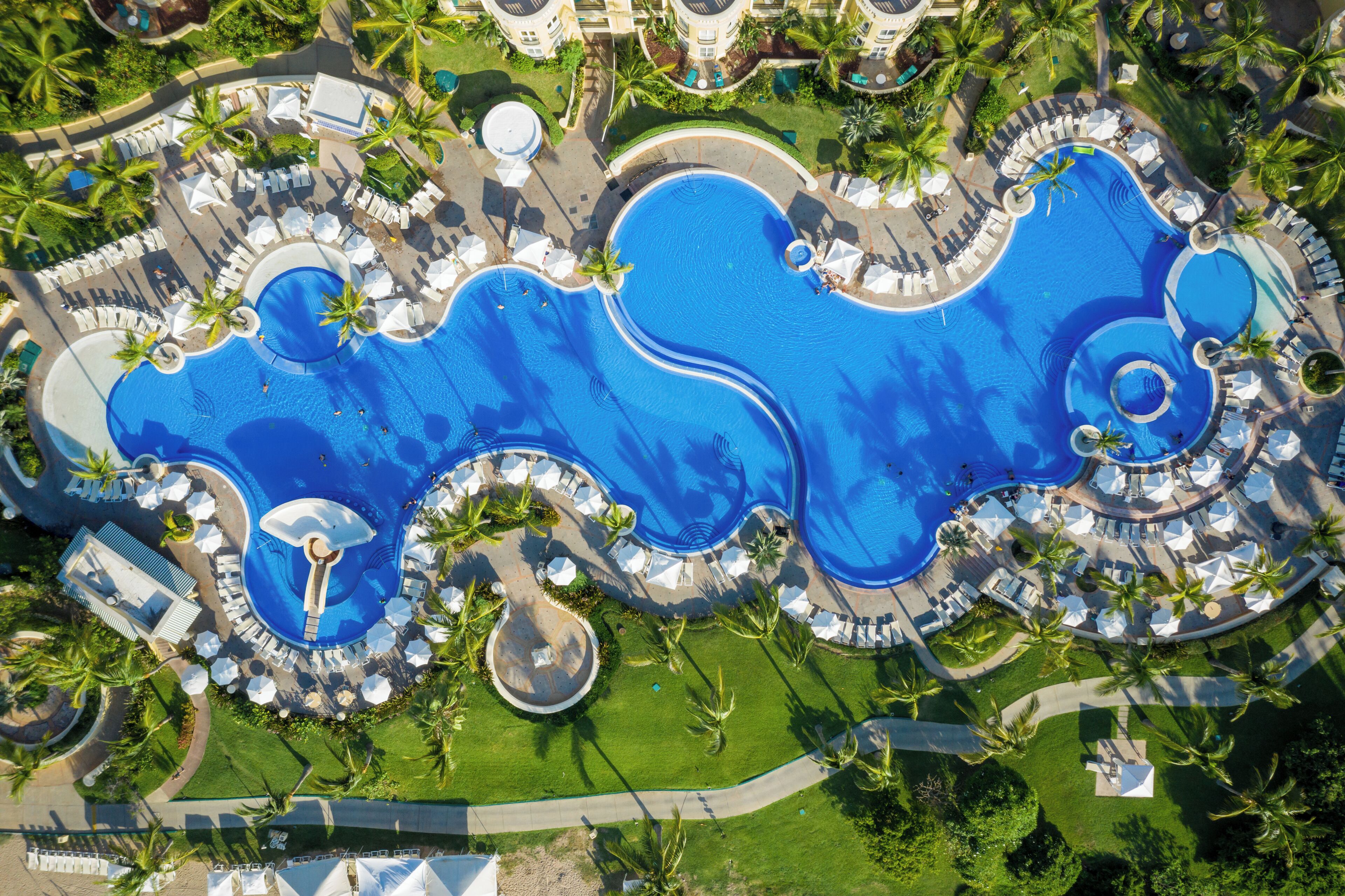 Vista aérea de un complejo turístico con varias piscinas, sillas de sol y palmeras.