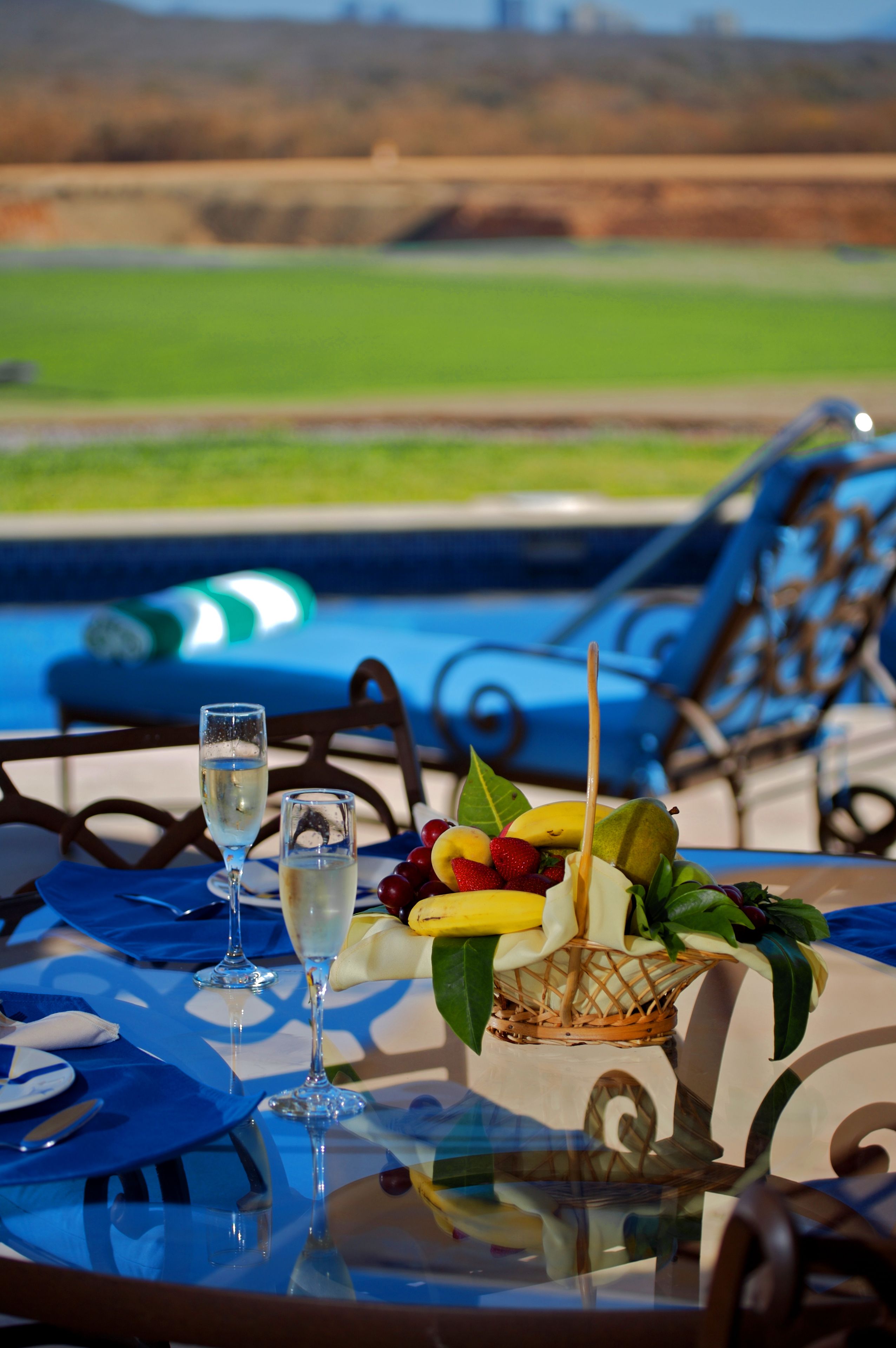 A table with a glass of water and a glass of wine, a fruit basket, and a view of a swimming pool and chairs.
