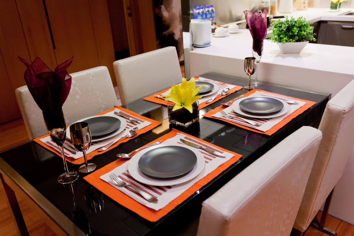A dining table set with plates, cutlery, and wine glasses, with orange placemats and a vase with a yellow flower.