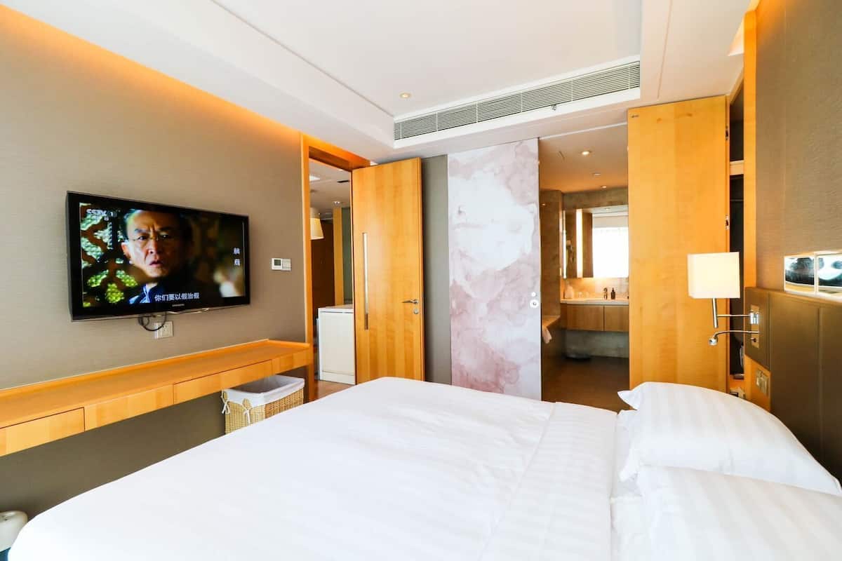 A hotel room with a large bed, a television mounted on the wall, a wooden desk, and a bathroom visible through an open door.