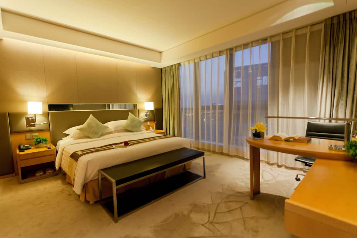 A hotel room with a bed, bedside tables, a desk, a bench, a TV, and a large window with curtains.