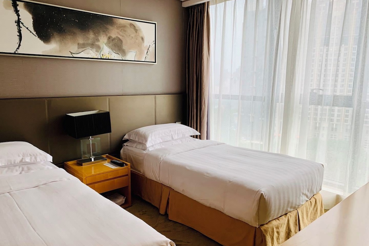 A hotel room with two beds, a nightstand, a lamp, a painting on the wall, and a window with curtains.