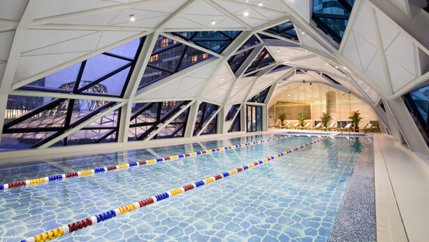 Una piscina interior con marcas divisorias claras, un diseño arquitectónico moderno con patrones geométricos y grandes ventanales que ofrecen vistas del paisaje urbano circundante.