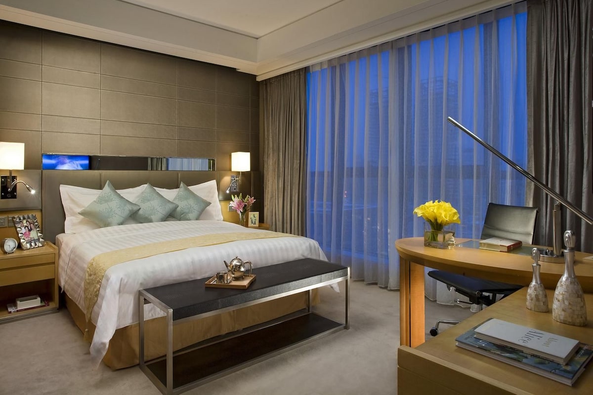 A hotel room with a bed, bedside tables, a desk, a chair, a TV, and a large window with curtains.