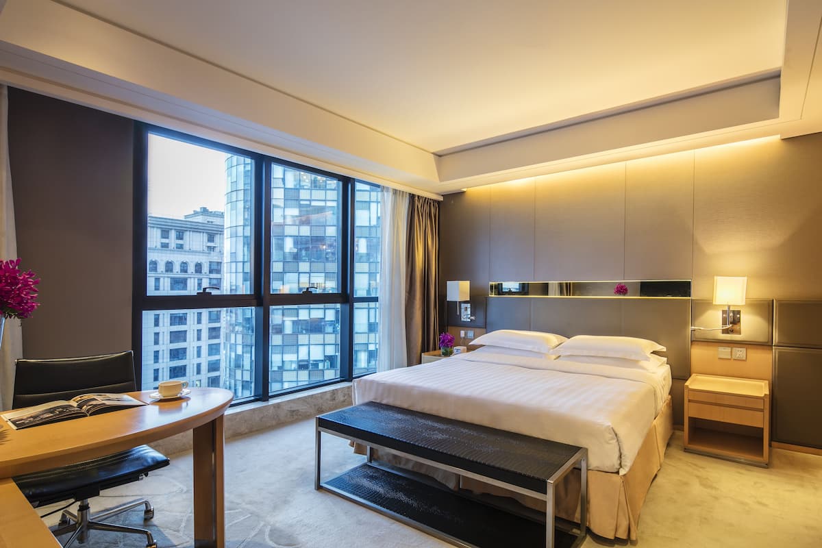 A hotel room with a large window, a bed, a desk, and a bench.