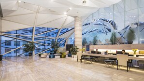 Un vestíbulo amplio con diseño moderno, que destaca por un techo alto con motivos geométricos, ventanas grandes y un mostrador de recepción con asientos.