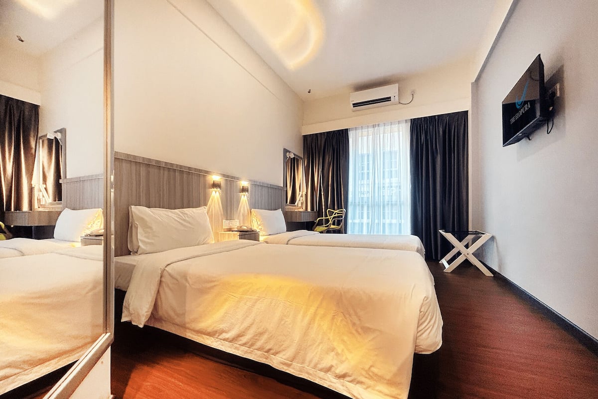 A hotel room with two beds, a flat-screen TV mounted on the wall, an air conditioning unit, and a window with curtains.