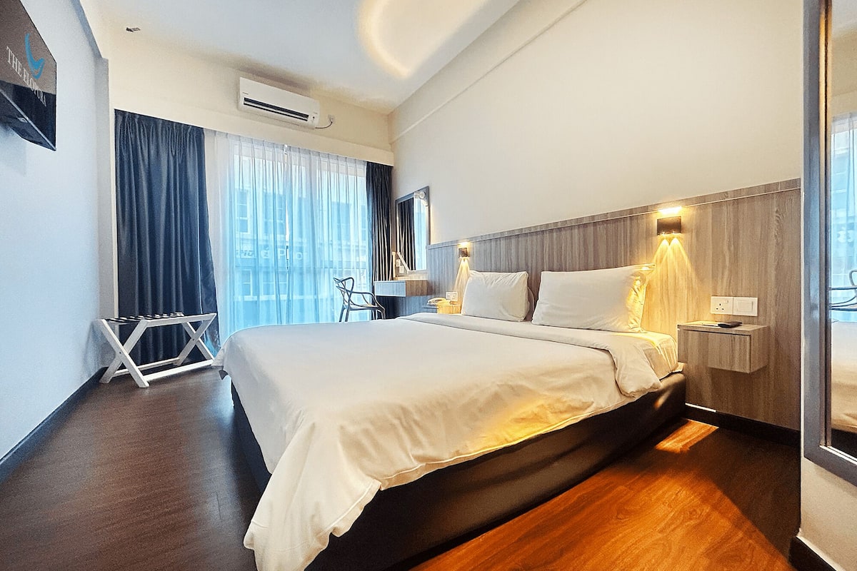 A modern hotel room with a large bed, a wooden floor, a desk with a chair, and a window with curtains.