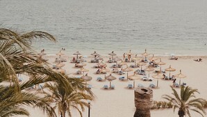 Una playa con filas de tumbonas y sombrillas de paja, palmeras y un mar tranquilo con montañas a lo lejos.