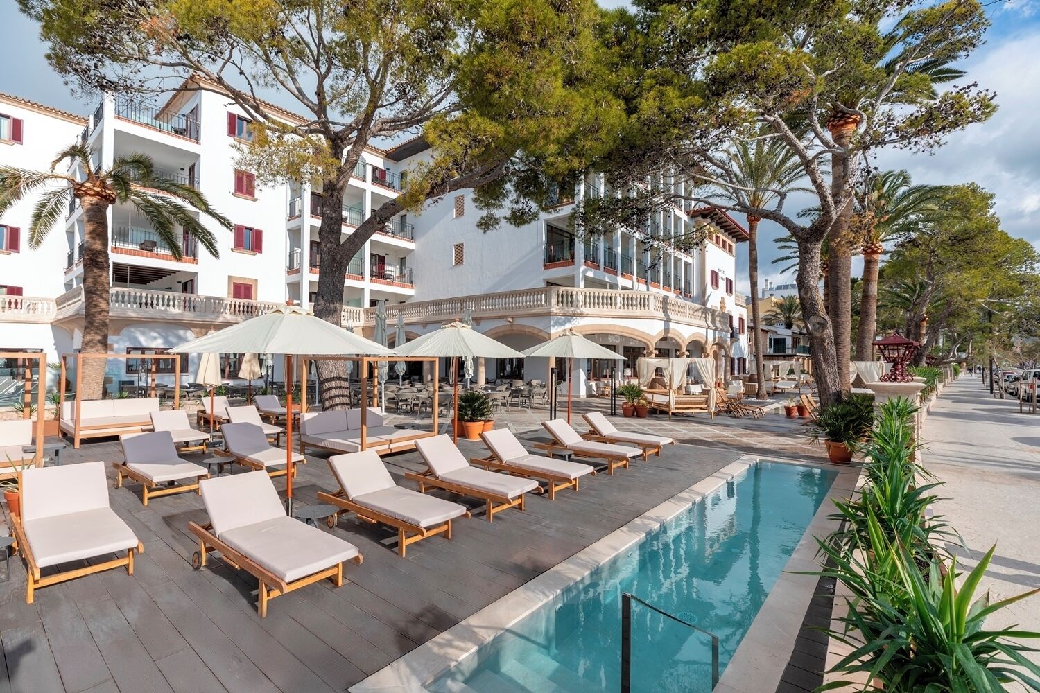 Uma área à beira da piscina com espreguiçadeiras e guarda-sóis, cercada por árvores e um edifício com varandas.