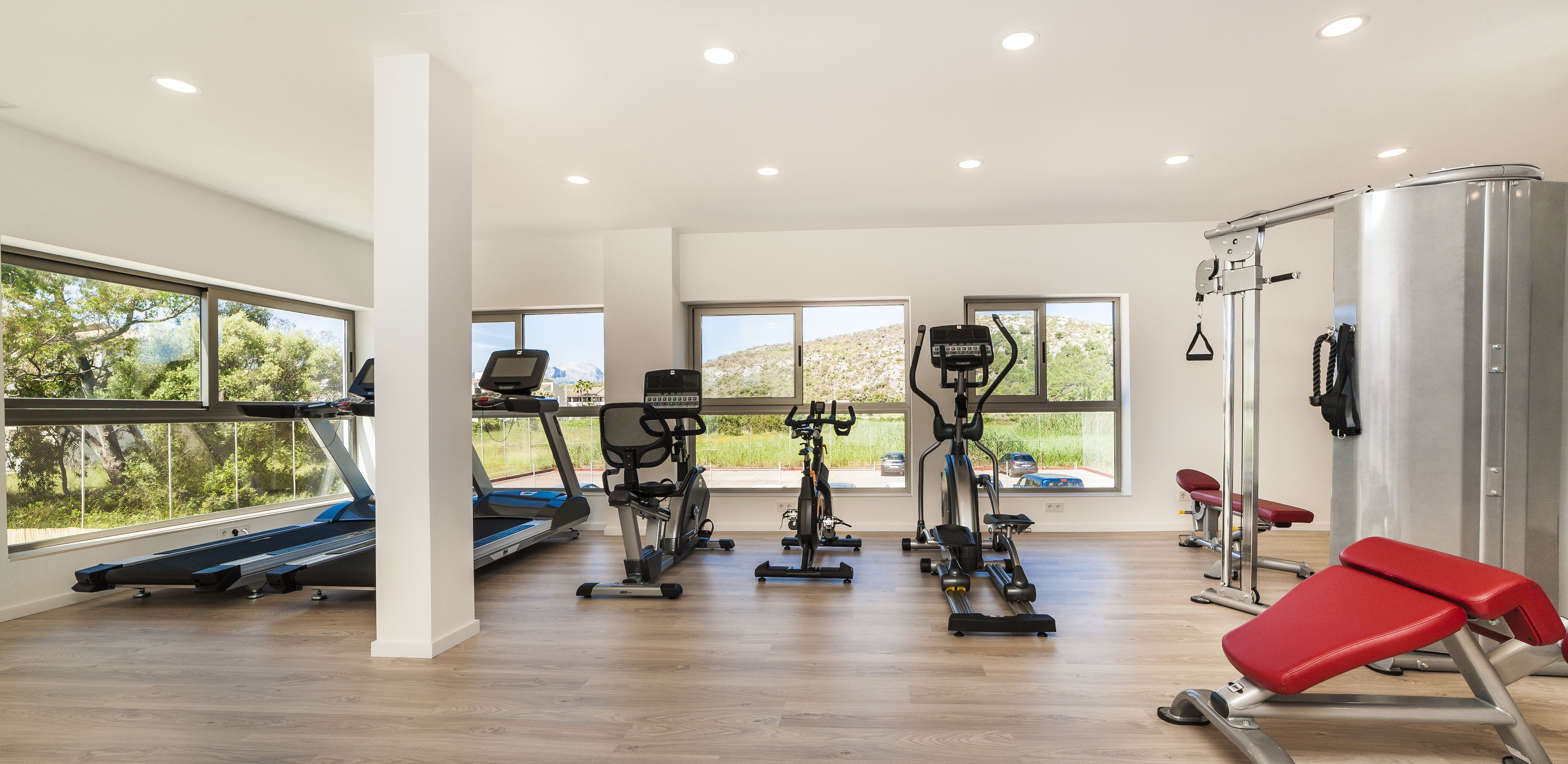 A modern gym with cardio machines, weightlifting equipment, and large windows offering views of greenery.