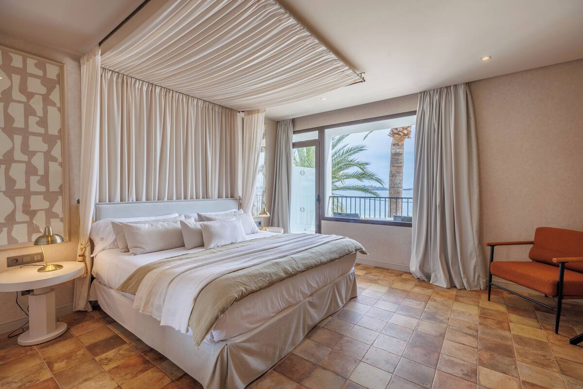 Quarto de hotel moderno com uma cama grande, área de estar, vista para palmeiras e uma varanda.