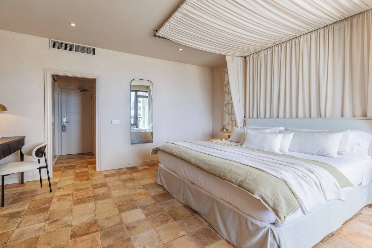 Quarto de hotel com uma cama grande, uma escrivaninha com cadeira, um espelho e uma porta que dá para o banheiro.