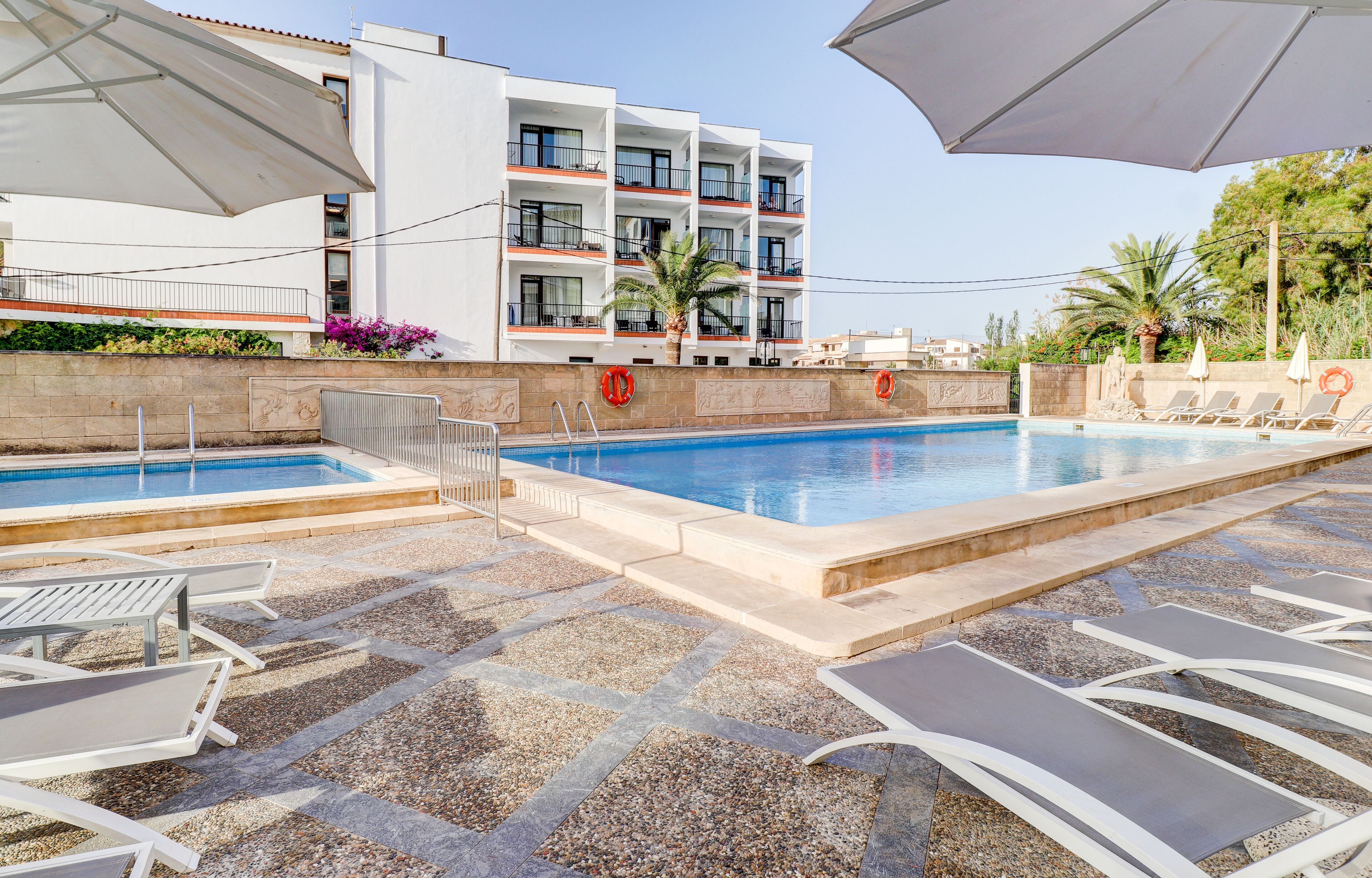 Zona de piscina hotelera con sillones de descanso y sombrillas, una piscina azul cristalina y un edificio con balcones.