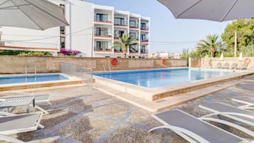 Zona de piscina hotelera con sillones de descanso y sombrillas, una piscina azul cristalina y un edificio con balcones.