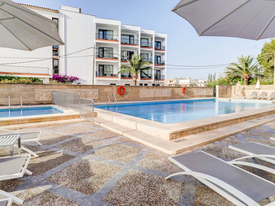 Zona de piscina hotelera con sillones de descanso y sombrillas, una piscina azul cristalina y un edificio con balcones.
