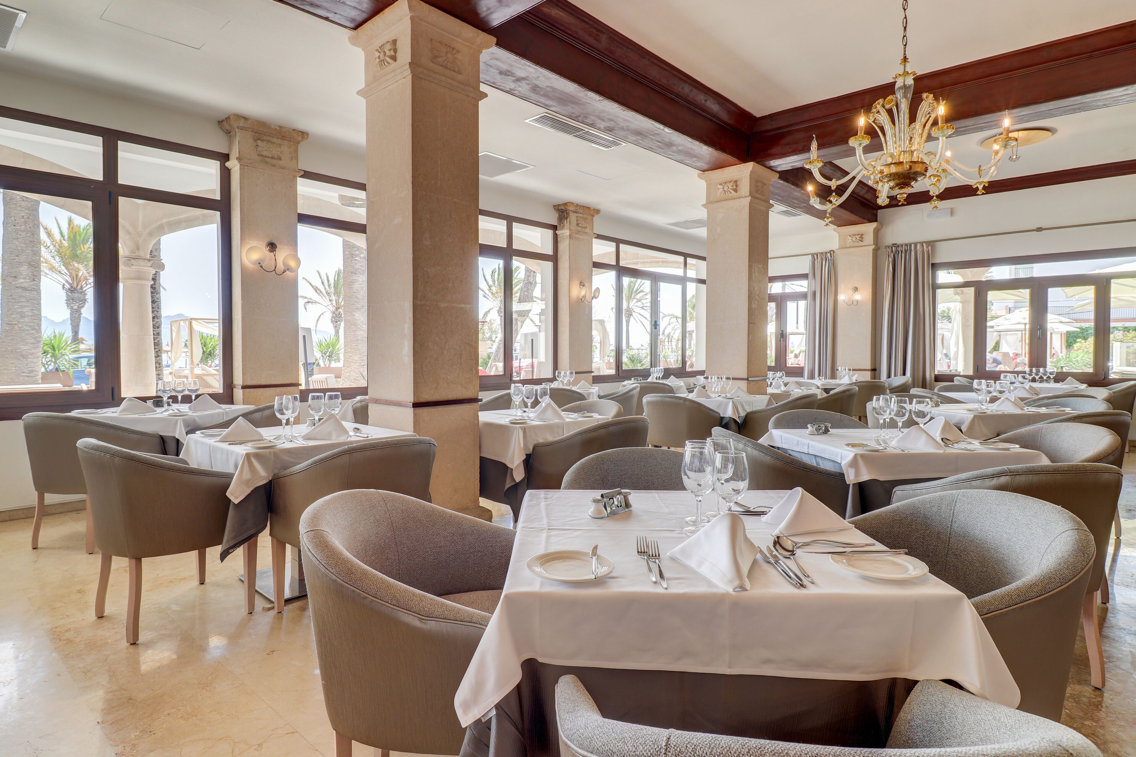A spacious dining area with multiple tables set for dining, large windows offering views of palm trees, and a chandelier hanging from the ceiling.